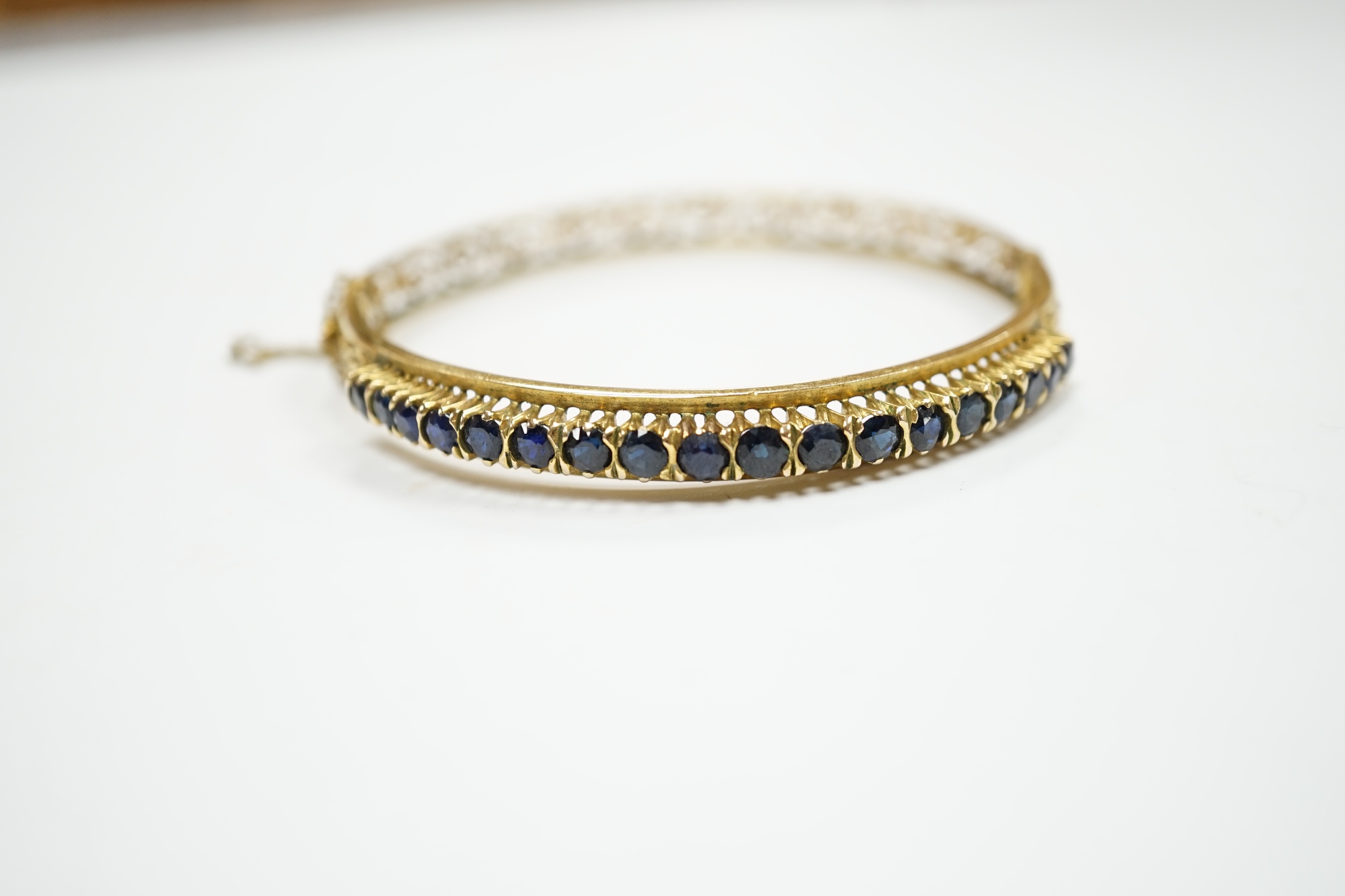 A pierced yellow metal and eighteen stone sapphire set hinged bangle, interior diameter 58mm, gross weight 14.1 grams.